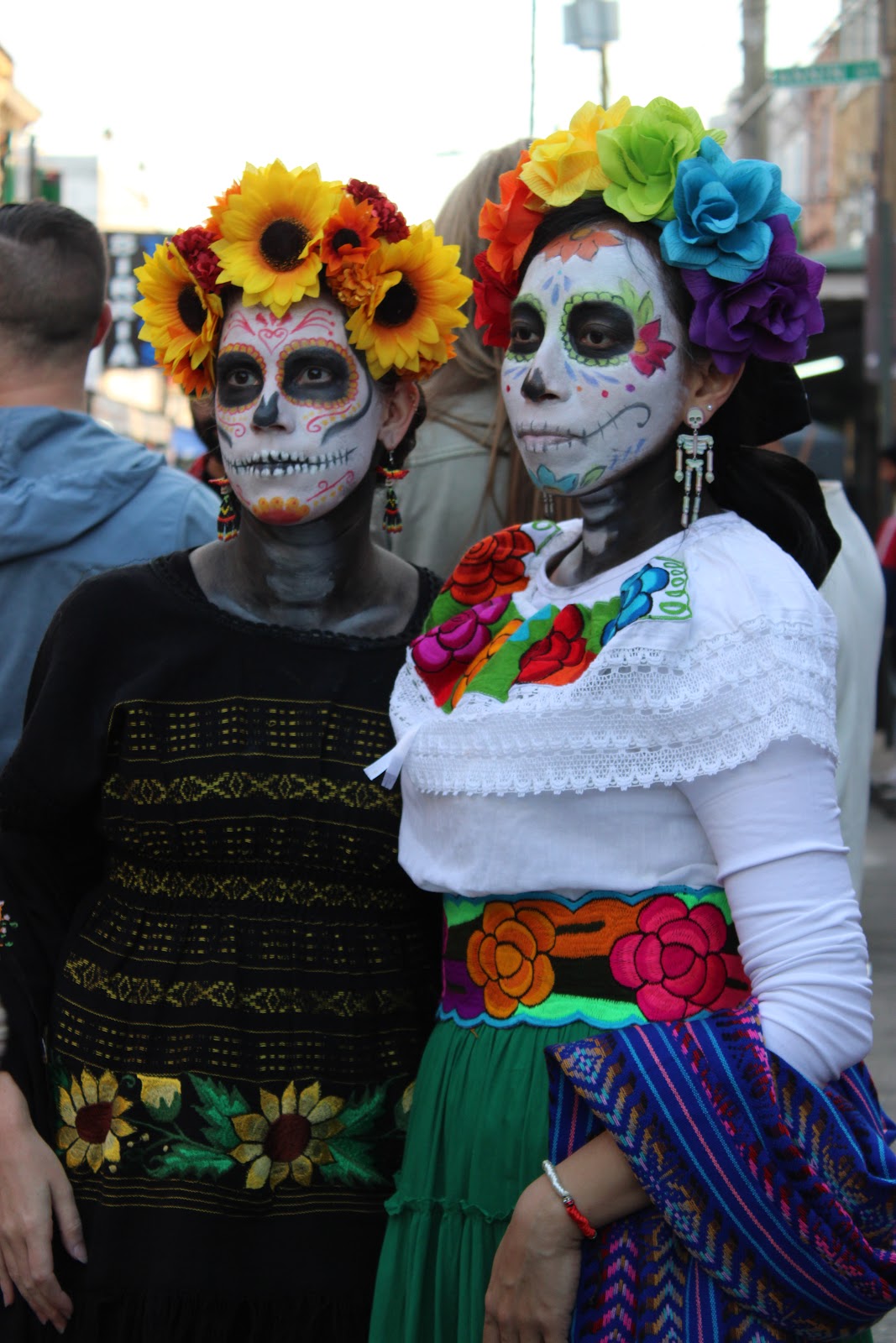 Two women in embroidered dresses, floral headdresses and Catrina face paint
