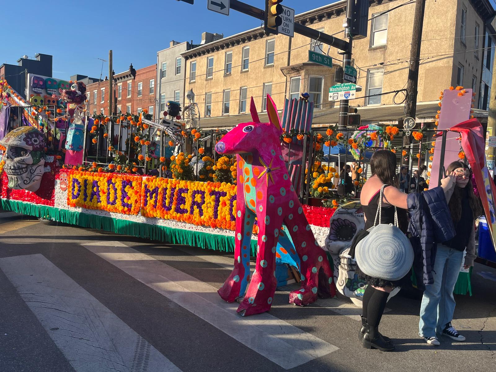 Large pink alebrije sculpture beside a Día de Muertos parade float on 9th Street